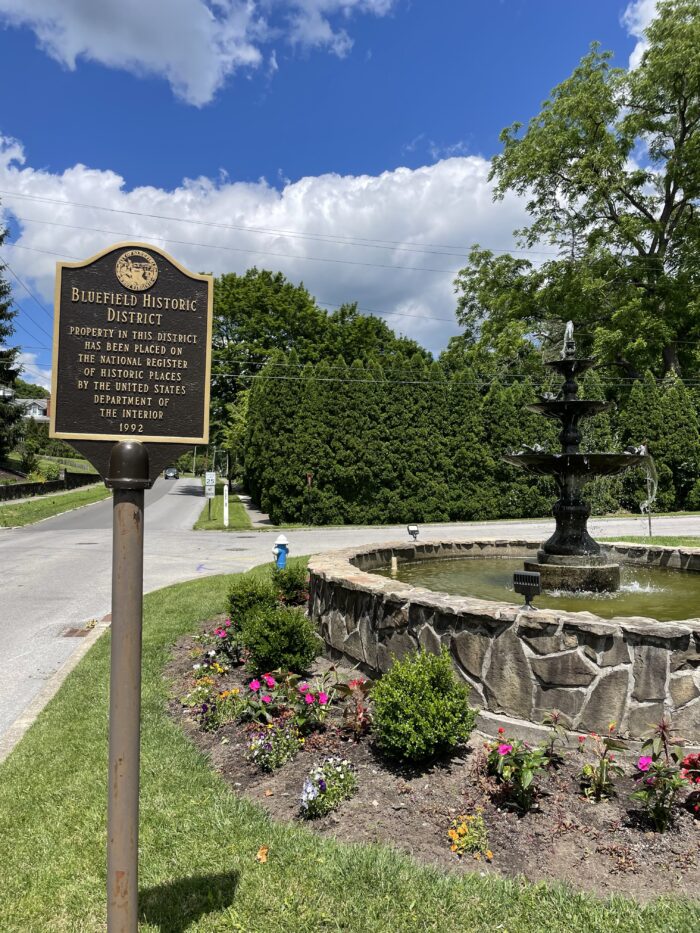 Historical marker sign and stone fountain surrounded by flowers at the start of the Bluefield Historical Home Driving Tour, with trees and a blue sky in the background.
