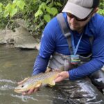 Man in fishing gear holding a brown trout in a stream, promoting West Virginia Fly Fishing.