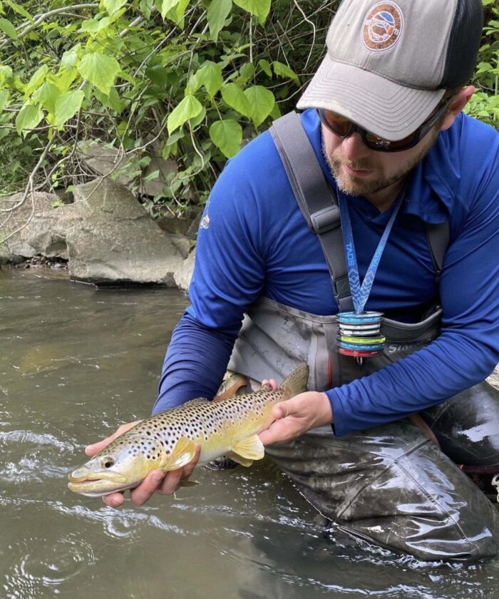 Man in fishing gear holding a brown trout in a stream, promoting West Virginia Fly Fishing.