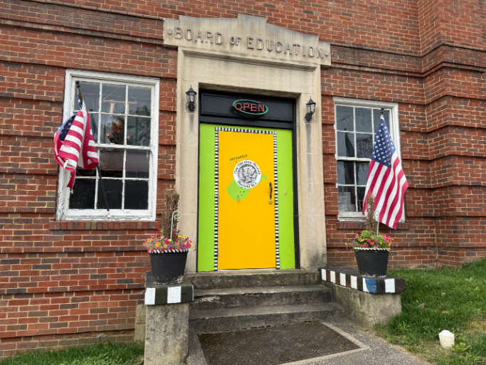 Entrance to the One Thin Dime Museum in a historic brick building with a bright yellow and green door, two American flags displayed on either side, and flower planters with black and white checkered bases.