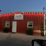Front view of Travis Collis Designs, a red metal building with a white door, two small windows, and potted plants out front. String lights line the roof and create a glowing effect against the evening.