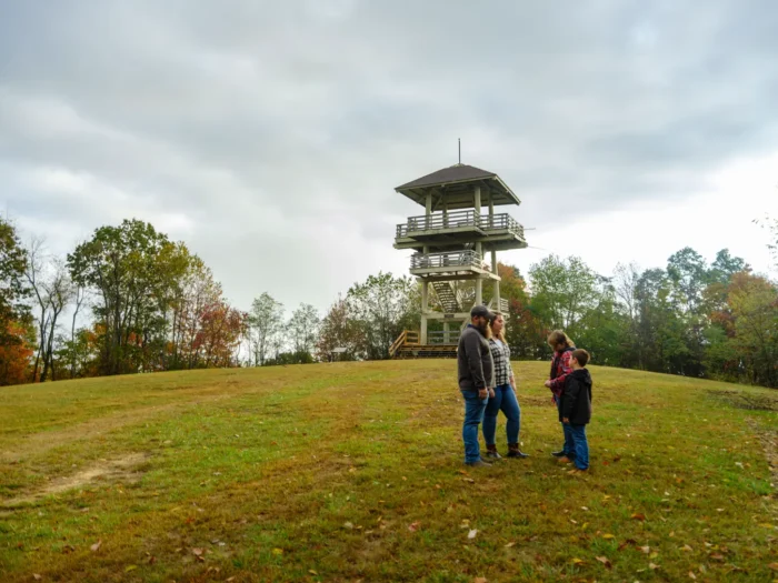 A family of four stands on a grassy hill at Pipestem Resort State Park, with the wooden Bolar Lookout Tower rising behind them and fall foliage starting to show in the surrounding trees.