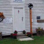 Todd Flat Historical School entrance with a white door, school bell on a post, and a sign beside the doorway on the white wooden exterior.