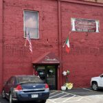 Exterior of Portabella Italian Restaurant in a historic red brick building. A maroon awning marks the entrance, with American and Italian flags mounted above the doorway. A large restaurant sign is displayed on the upper wall. Two vehicles are parked in front, and flower arrangements and balloons decorate the entrance. White parking lines, including an accessible space, are visible in the foreground.