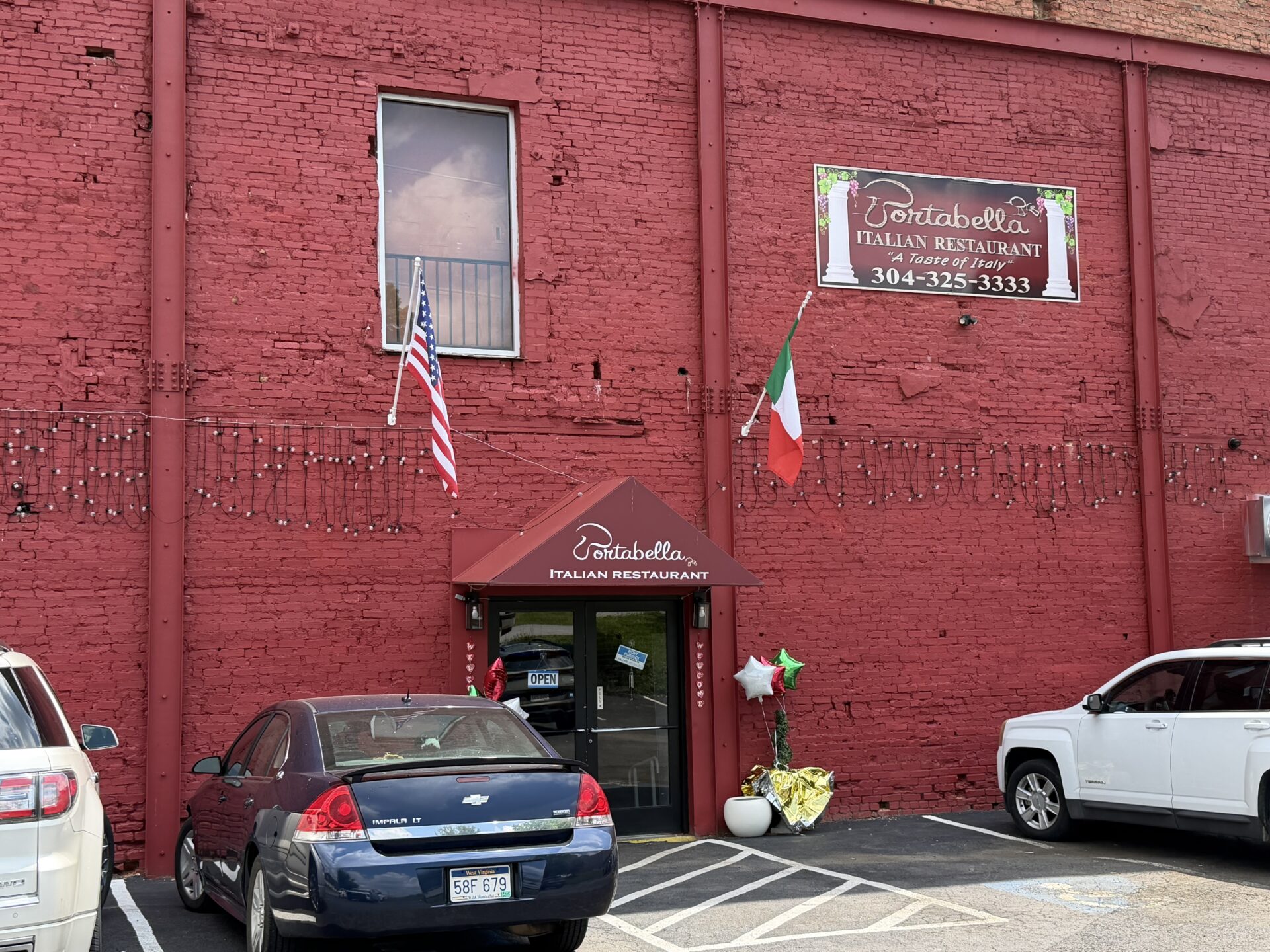 Exterior of Portabella Italian Restaurant in a historic red brick building. A maroon awning marks the entrance, with American and Italian flags mounted above the doorway. A large restaurant sign is displayed on the upper wall. Two vehicles are parked in front, and flower arrangements and balloons decorate the entrance. White parking lines, including an accessible space, are visible in the foreground.