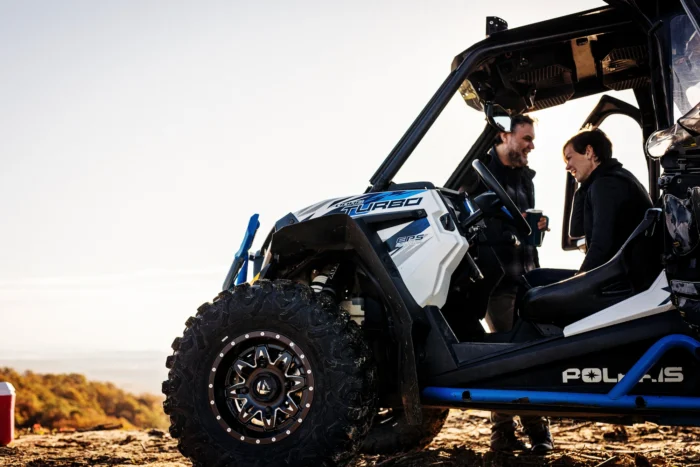 Close-up of a Polaris Turbo off-road vehicle (UTV) with two people sitting inside, engaged in friendly conversation. The driver and passenger are smiling at each other against a bright, clear sky backdrop. The vehicle features a white and blue color scheme with prominent Polaris branding. Its large, rugged tire with a decorative rim is visible in the foreground. The image appears to be taken at an elevated viewpoint during sunset or sunrise.