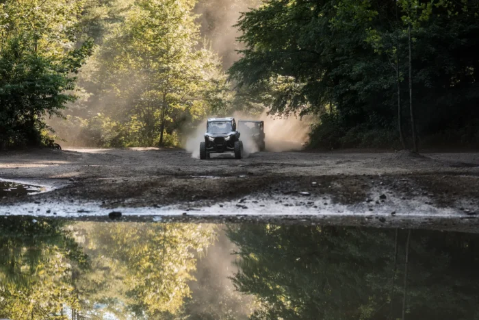 Two off-road vehicles driving down a dusty forest trail, kicking up clouds of dust that catch the sunlight filtering through the trees. The scene features lush green trees on both sides of the dirt path, with dappled sunlight creating a beautiful atmosphere. At the bottom of the image is a puddle that perfectly mirrors the forest and vehicles, creating a symmetrical reflection. The overall mood conveys adventure and exploration in a serene natural setting.