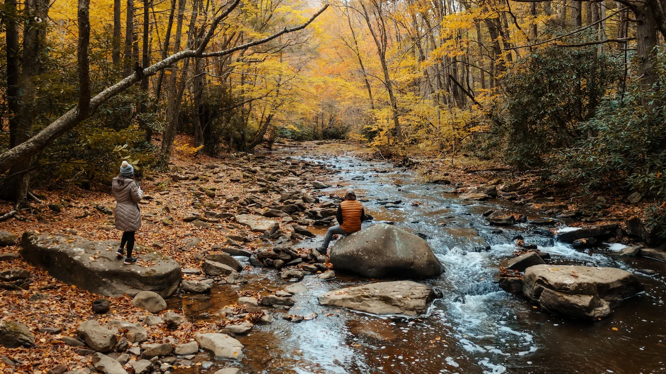 A couple in comfortable fall apparel rest and admire a gently rolling stream, surrounded by brightly colored fall foliage