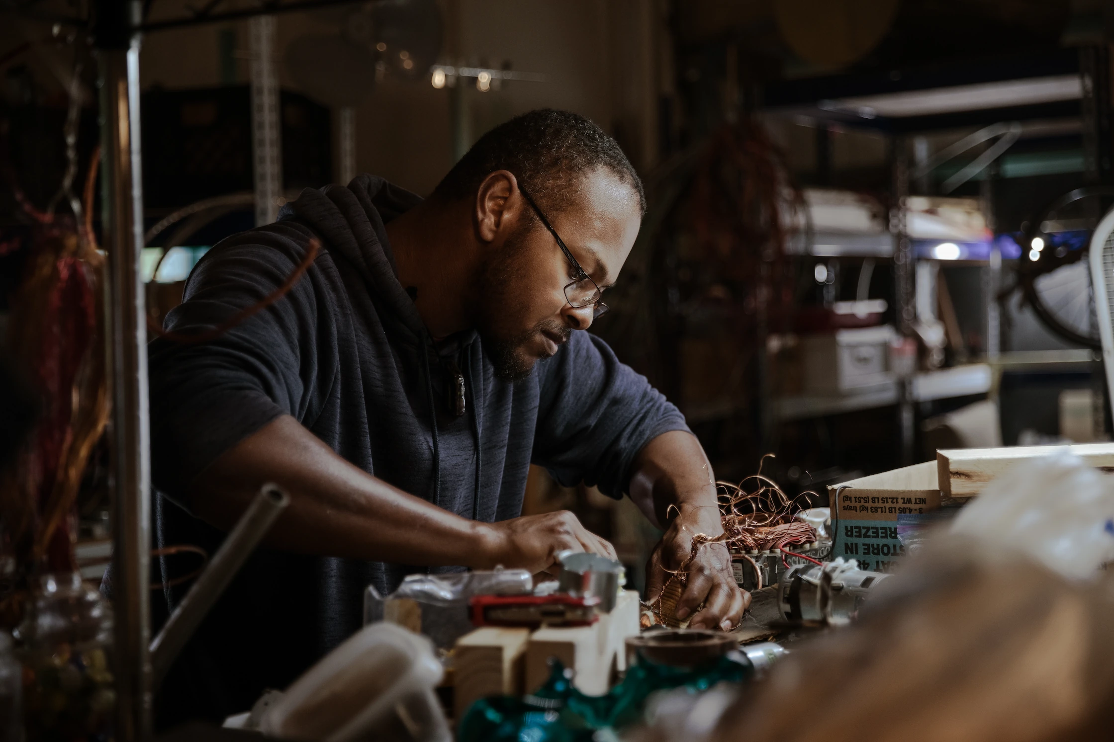 A black male artist with glasses looks thoughtfully at a wire sculpture he is constructing in his workshop.