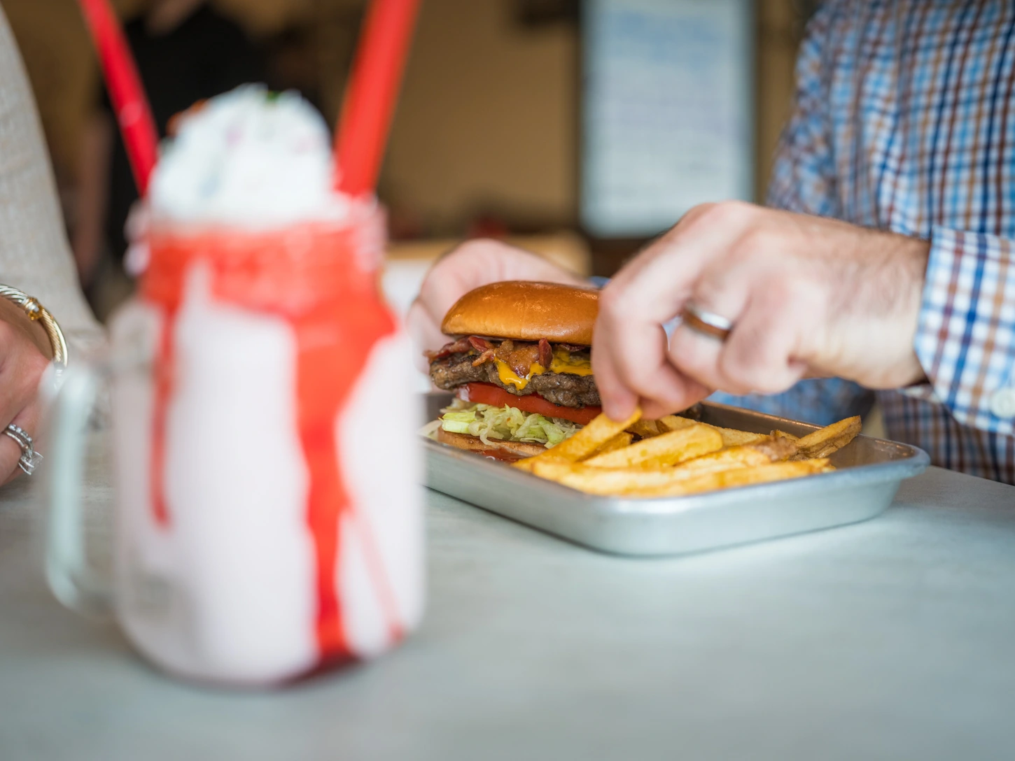 A metal food tray at Jimmie's Restaurant - containing an appetizing bacon cheeseburger with lettuce and tomato on a buttered and toasted bun and golden french fries. Nearby, there is a strawberry milkshake toped with whipped cream and sprinkles and a drizzle of strawberry syrup.