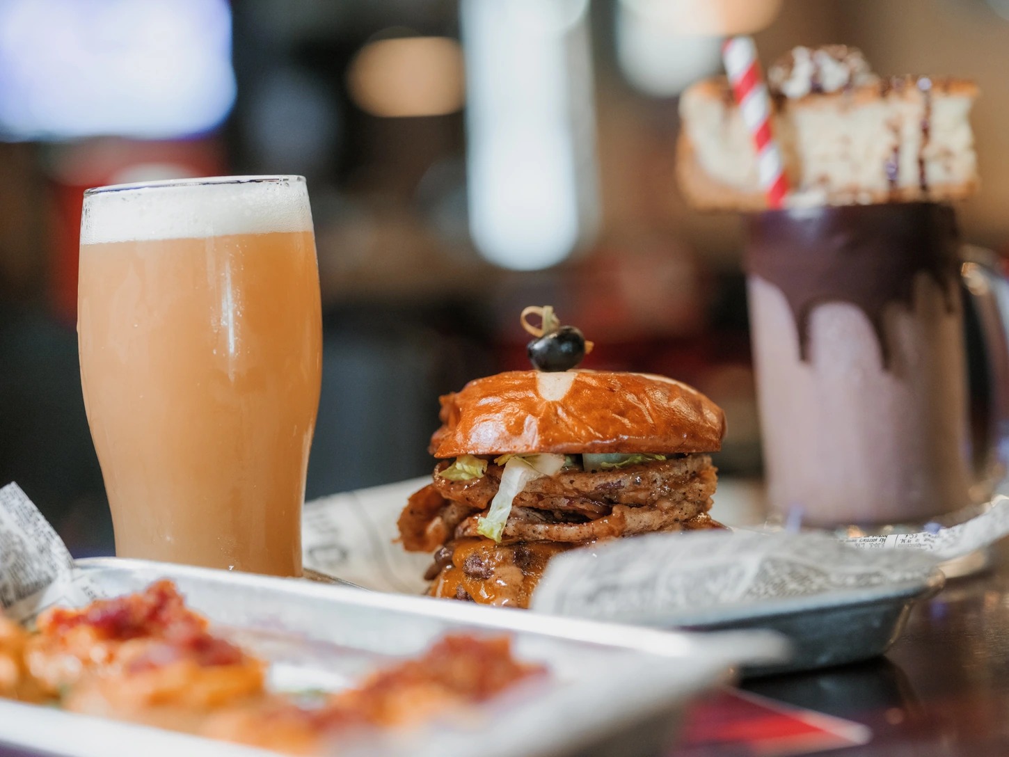 A mouth watering spread of food atop a bar at The Railyard. The spread includes a barbecue brisket sandwhich on a golden brioche bun, a tray of fried green tomatoes topped with bacon and pimento cheese, a milkshake drizzled with chocolate syrup and topped with a slice of cake, and a chilled pint glass of IPA