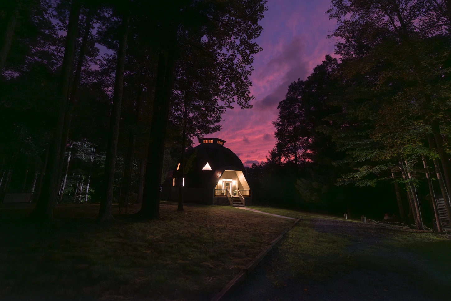 A geometric dome shaped AirBnB cabin. Lights from the windows and porch cast a gentle glow on the surrounding field. The cabin stands in stark contrast to the pink and purple hues of the evening sky