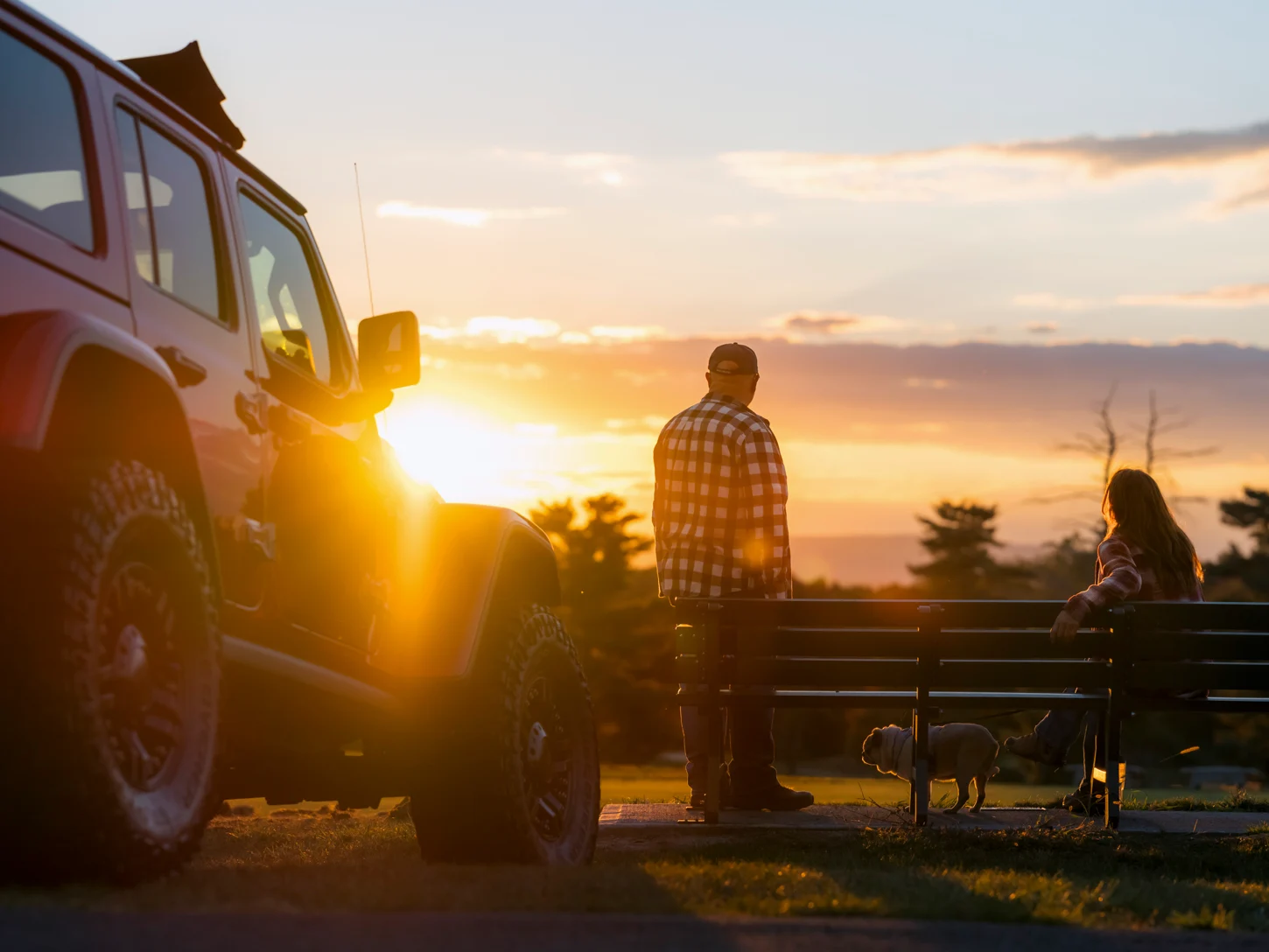 A couple watches a serene sunset from a park bench in Pipestem State park - the sunlight glints off the hood of their red jeep, and a small pug stands near their feet, on leash