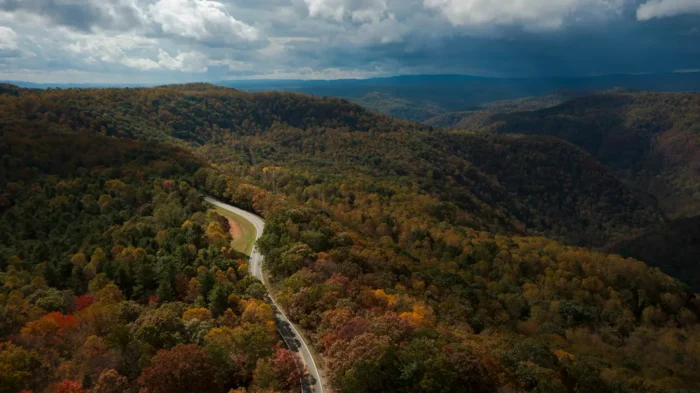 A scenic mountain road winds through the vibrant autumn forest of Mercer County, WV, with dramatic clouds casting shadows over the rolling Appalachian hills.