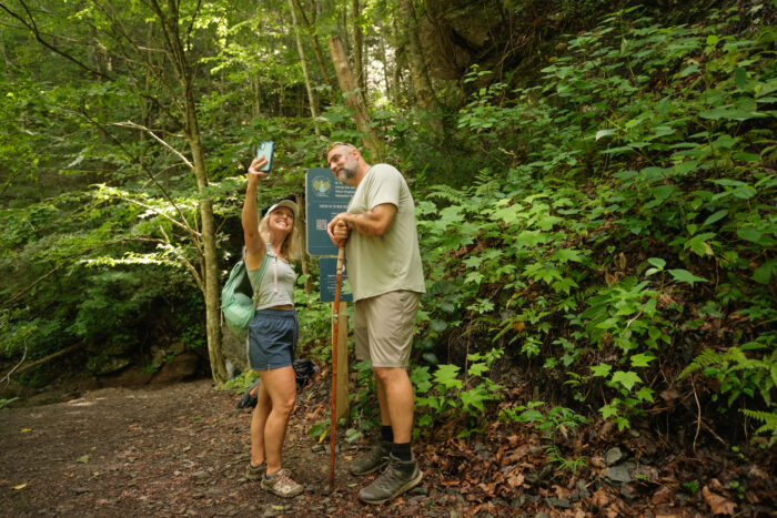 Two hikers pause on a forest trail to take a selfie next to a trail sign. The woman is smiling while holding up her phone, and the man leans on a wooden walking stick. Lush green foliage surrounds them.