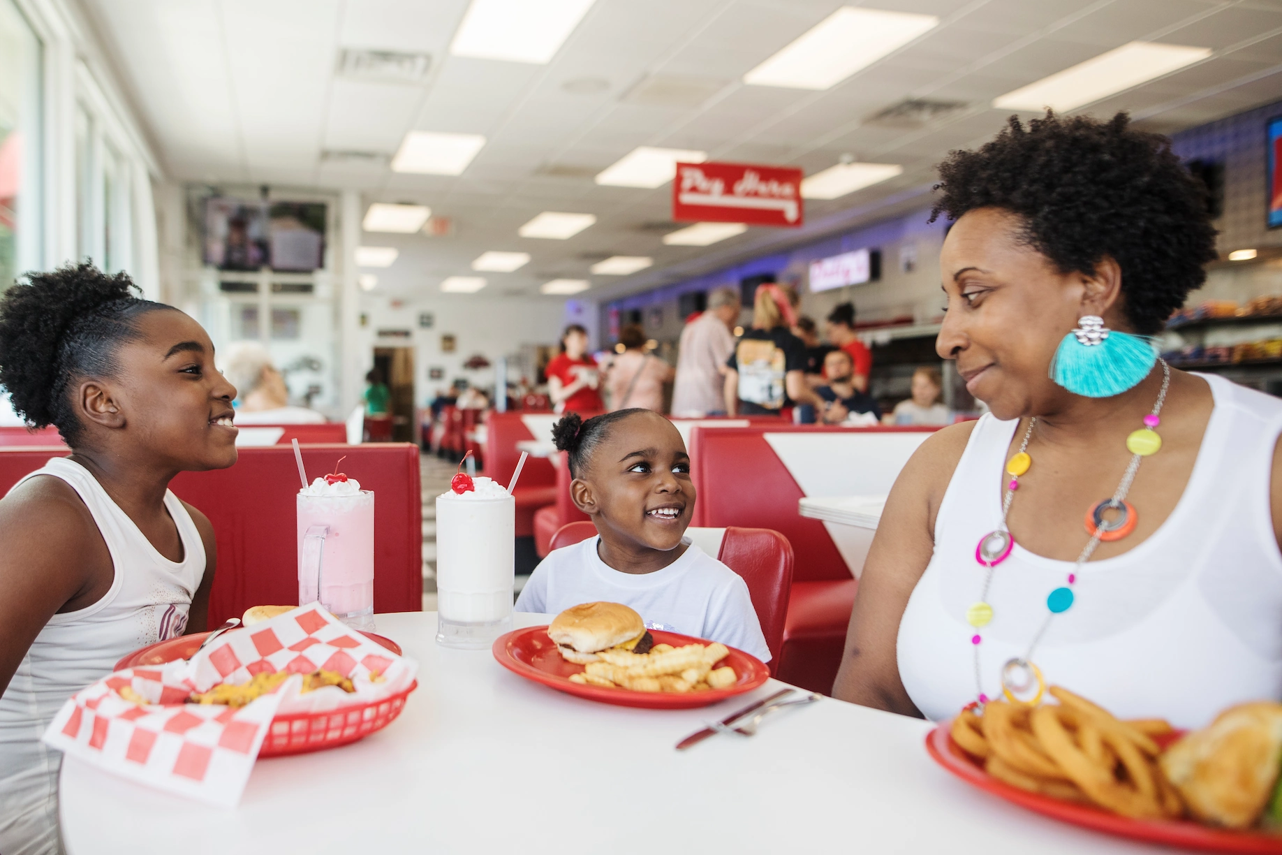 A mother and her two daughters seated at a booth in Dolly's Diner. Their table contains multiple plates of crispy onion rings, golden french fries, and classic cheeseburgers, and two milkshakes - each topped with whipped cream and a cherry