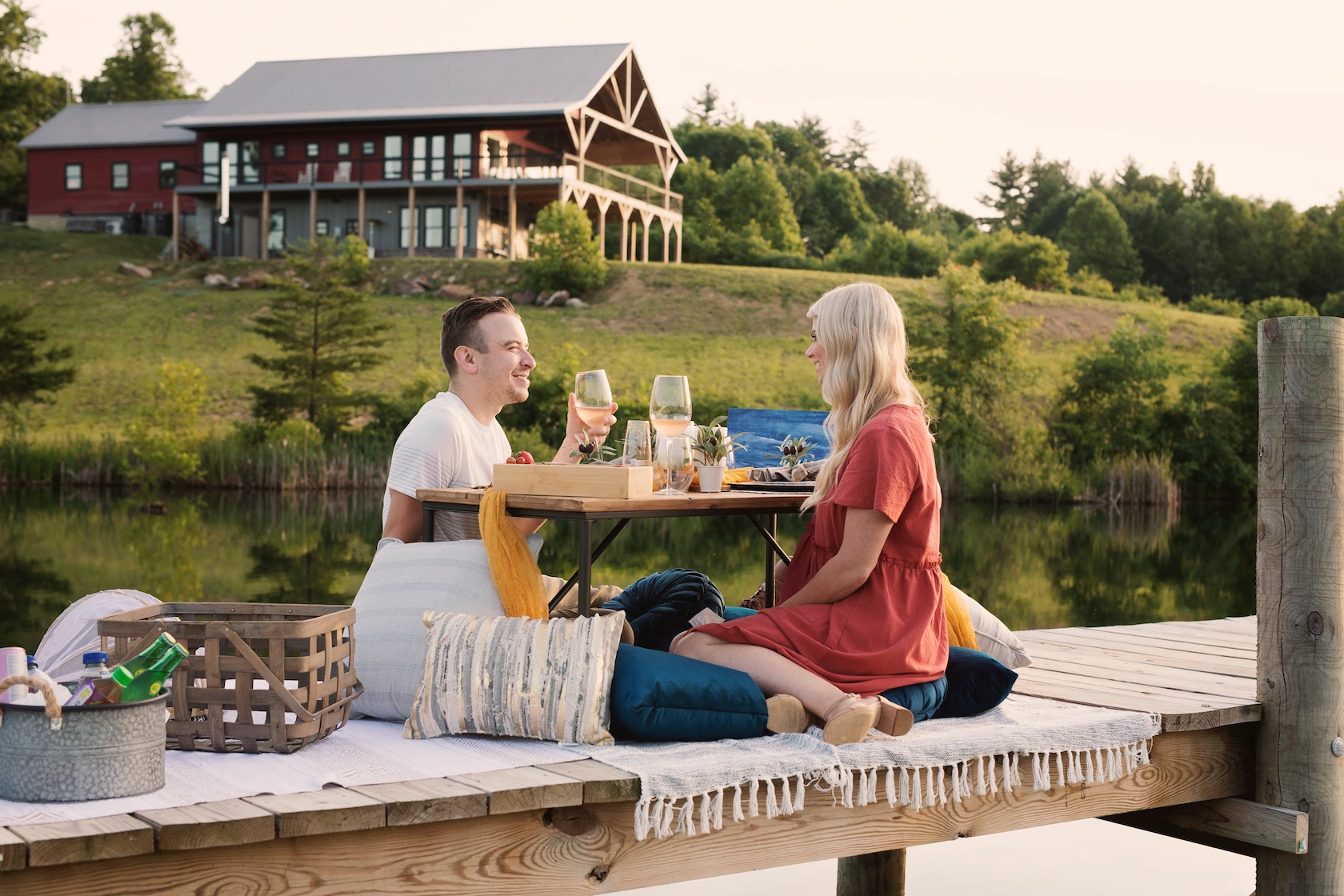 A young couple sips wine at a picnic on a wooden dock. A large red lake house, known as the Lake Mountain Manor, sits on a hill overlooking the lake.