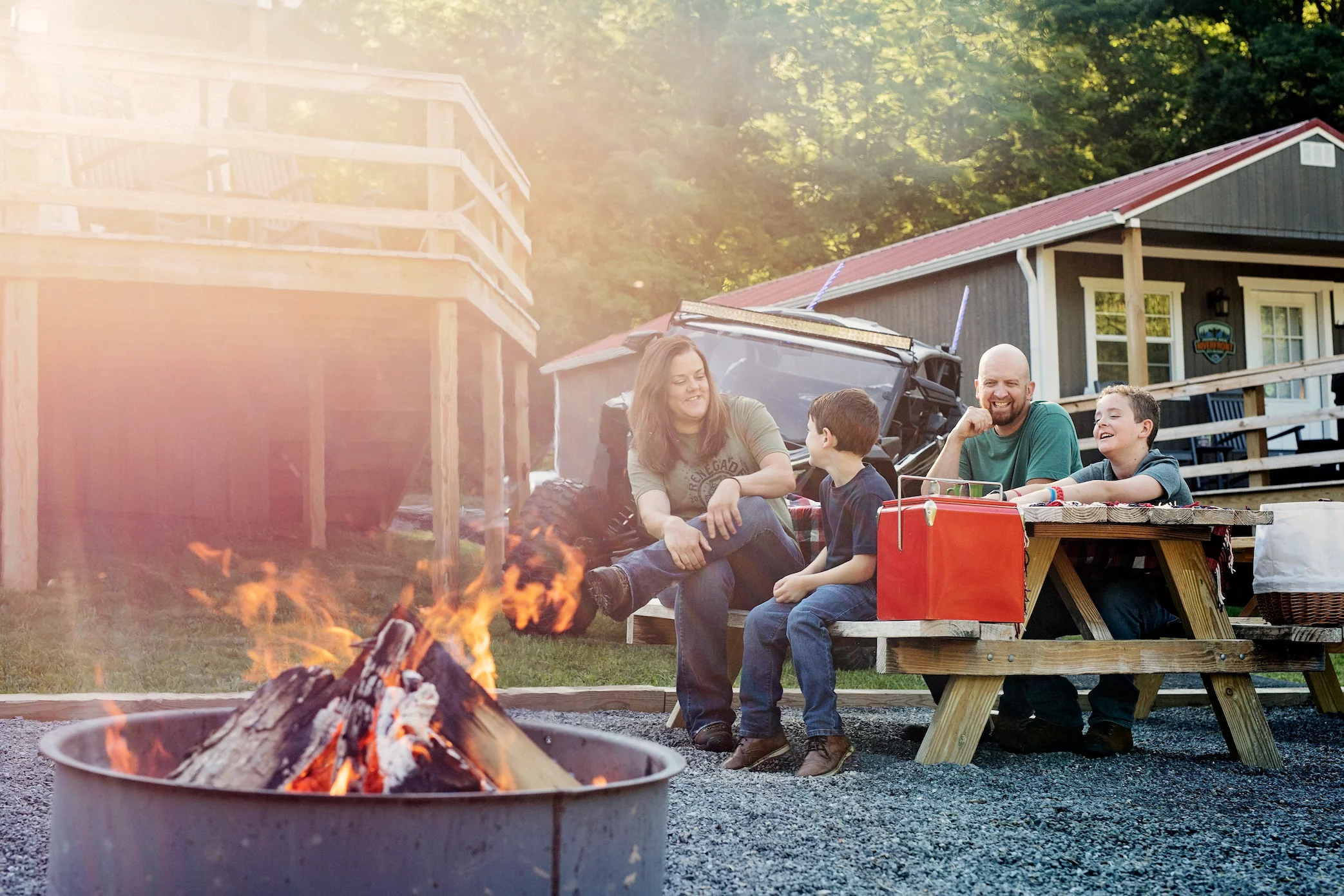 A family seated together at a picnic table next to a blazing campfire. An ATV and several cabins are visible in the background