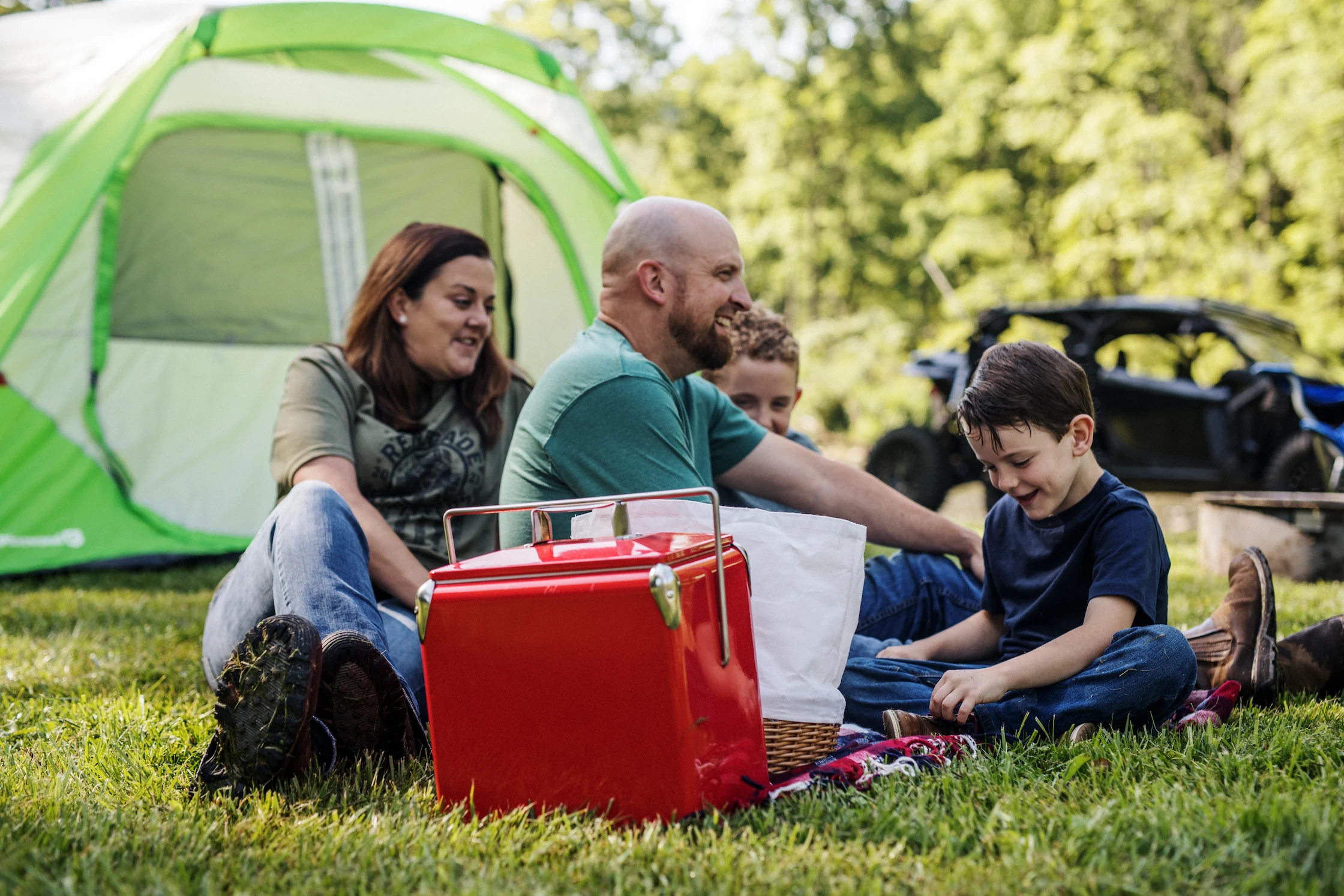 A family has a picnic and shares a laugh at their riverfront campsite. A green tent and ATV are visible in the background.