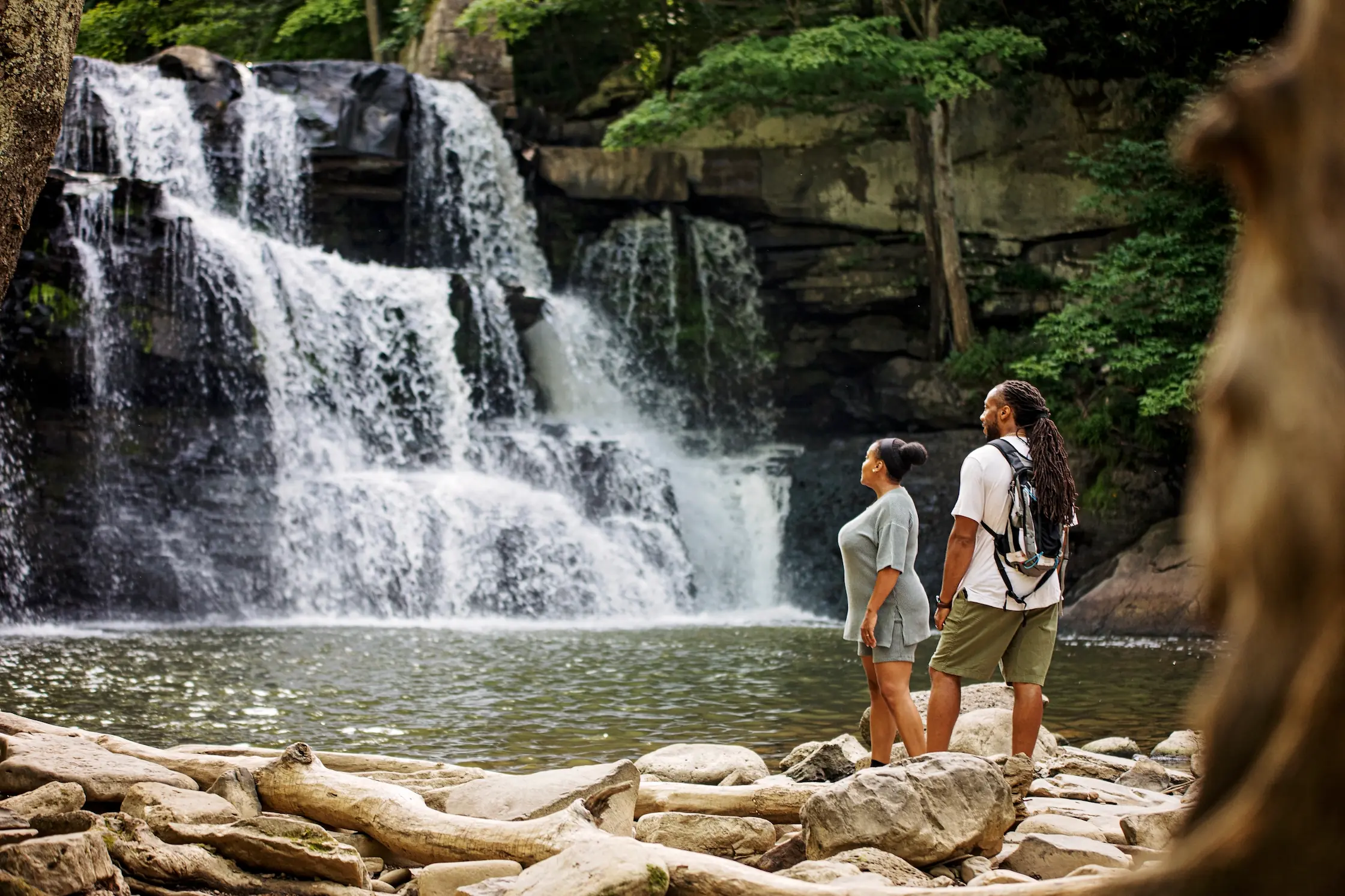 A couple with rich brown skin tones stand at the base of the Brush Creek waterfall and admire its beauty from the nearby shore.