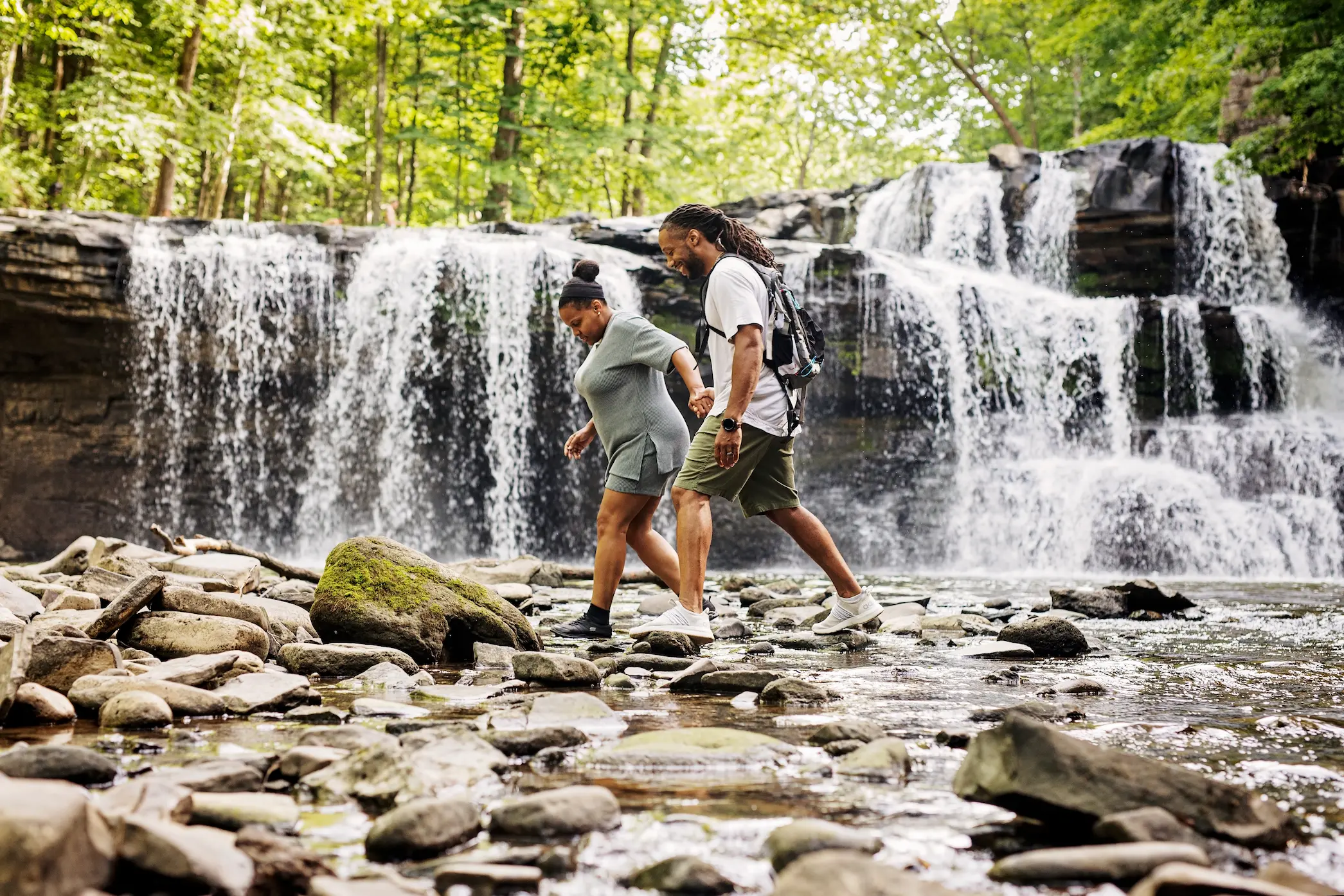 A couple with rich brown skin tones and casual athletic wear walk hand-in-hand across river stones in front of a gently cascading waterfall