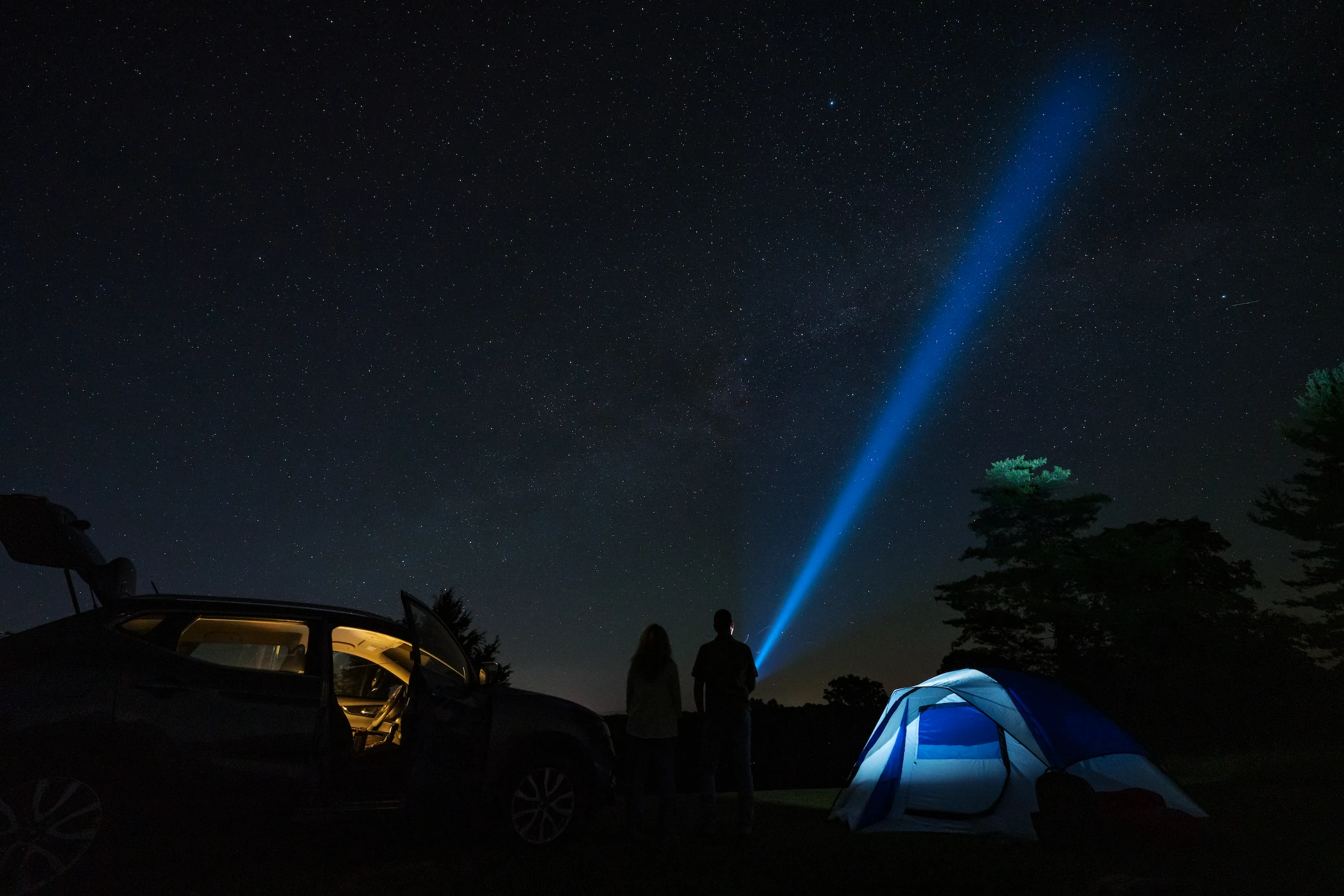 A couple shines a flashlight into the starry night sky. Light from their tent and an open car door cast a soft glow on the surrounding area.