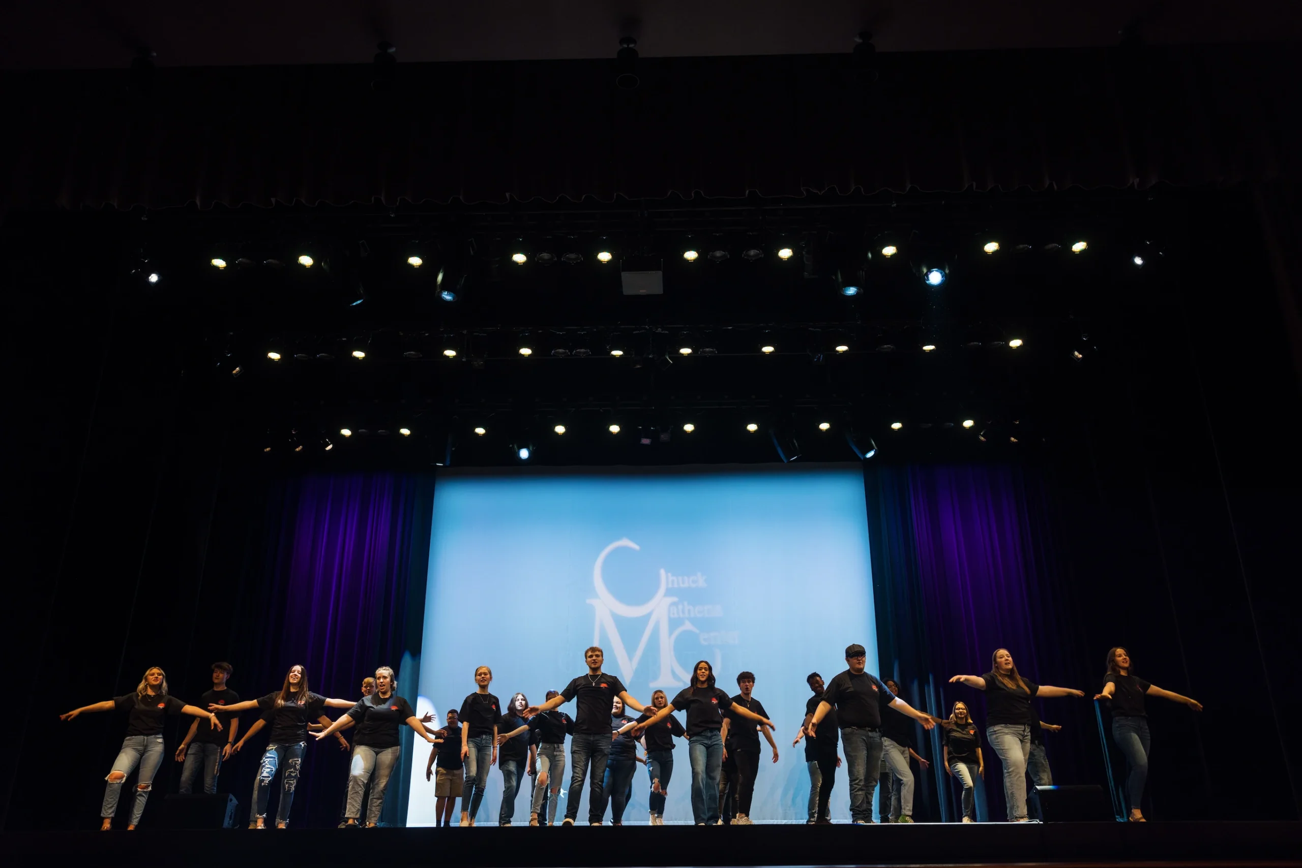 A diverse group of students perform in matching black t-shirts on the stage at the Chuck Mathena Center