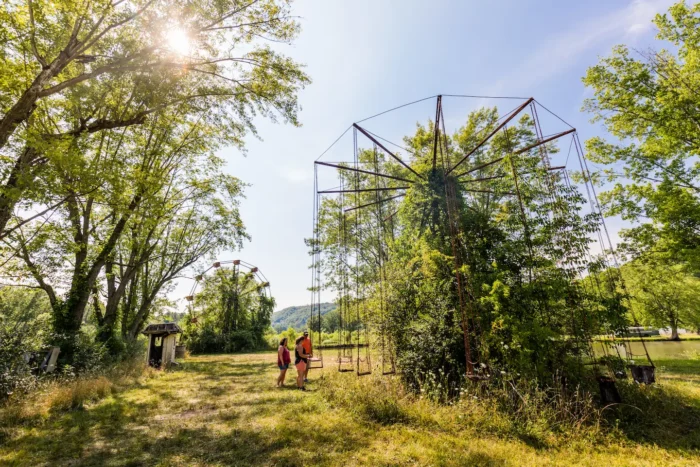 Two people explore an overgrown, abandoned amusement park with a rusted swing ride and a Ferris wheel partially hidden by trees under a bright, sunny sky.