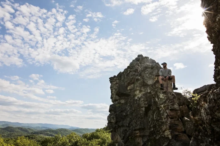 Man sitting on a rocky cliff overlooking forested hills under a bright sky.