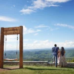 Three people standing near the Almost Heaven swing at East River Mountain Scenic Overlook in Mercer County, West Virginia, with sweeping views of green hills under a bright blue sky with clouds