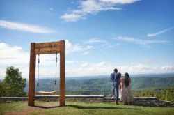 Three people standing near the Almost Heaven swing at East River Mountain Scenic Overlook in Mercer County, West Virginia, with sweeping views of green hills under a bright blue sky with clouds