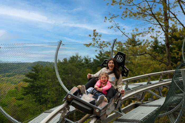 A mom and daughter rides the mountain coaster. The coaster is among the trees and the sky is blue.
