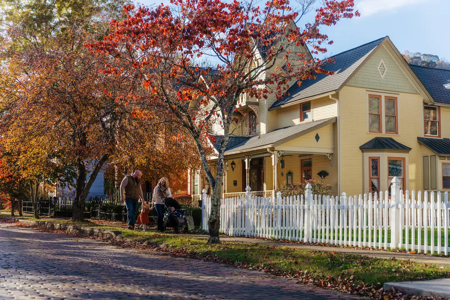 A family walks along a tree-lined brick street in Mercer County, WV, surrounded by vibrant fall foliage and historic homes with white picket fences.