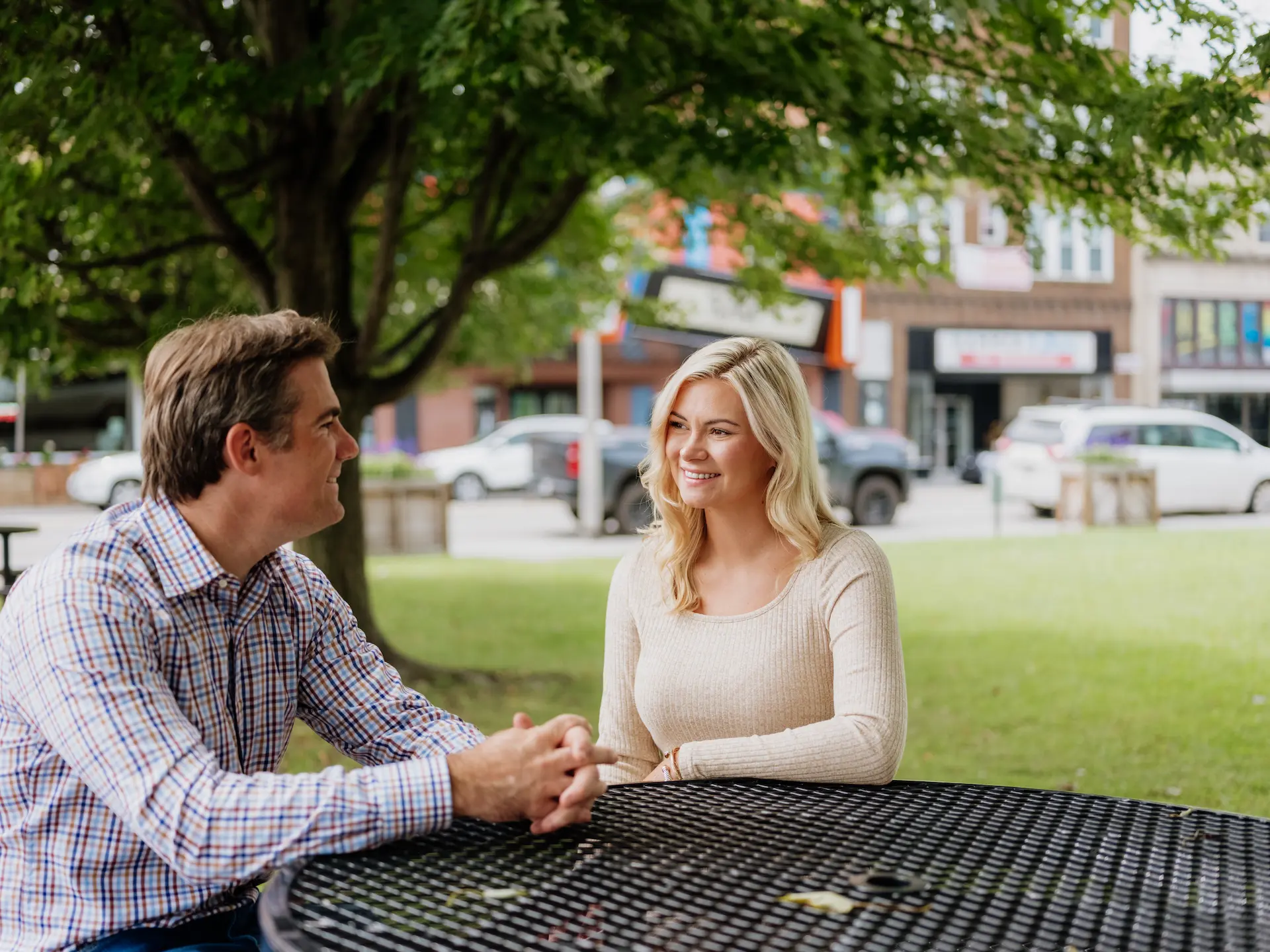 A couple sits at a black metal picnic table in downtown Mercer County, WV, enjoying a relaxed conversation beneath a leafy tree with local shops in the background.