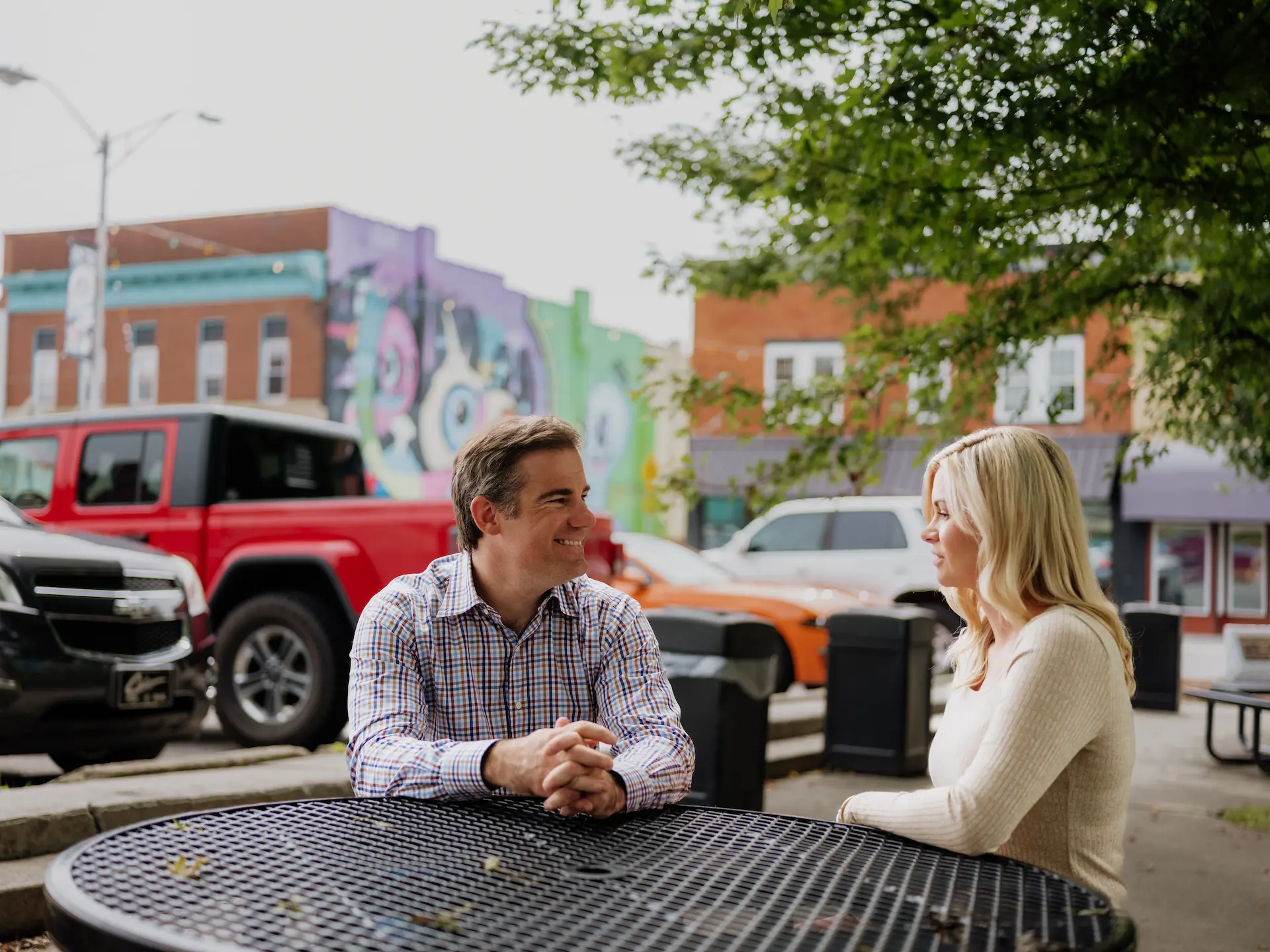 A couple sits at a black metal table in downtown Mercer County, WV, enjoying conversation with vibrant street murals and local storefronts in the background.