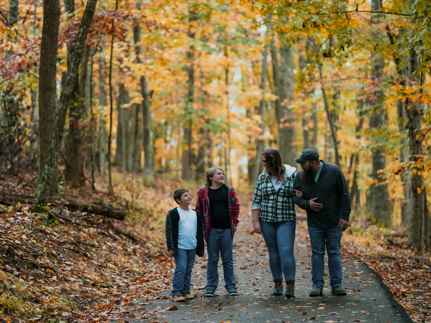 A family of four walks along a leaf-covered path in a forest with vibrant autumn foliage