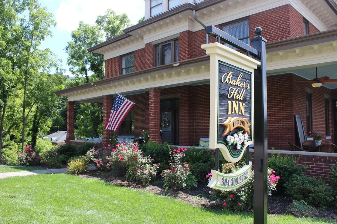 A red-brick bed and breakfast surrounded by flowers and greenery with an American flag and a sign reading Baker’s Hill Inn, circa 1910