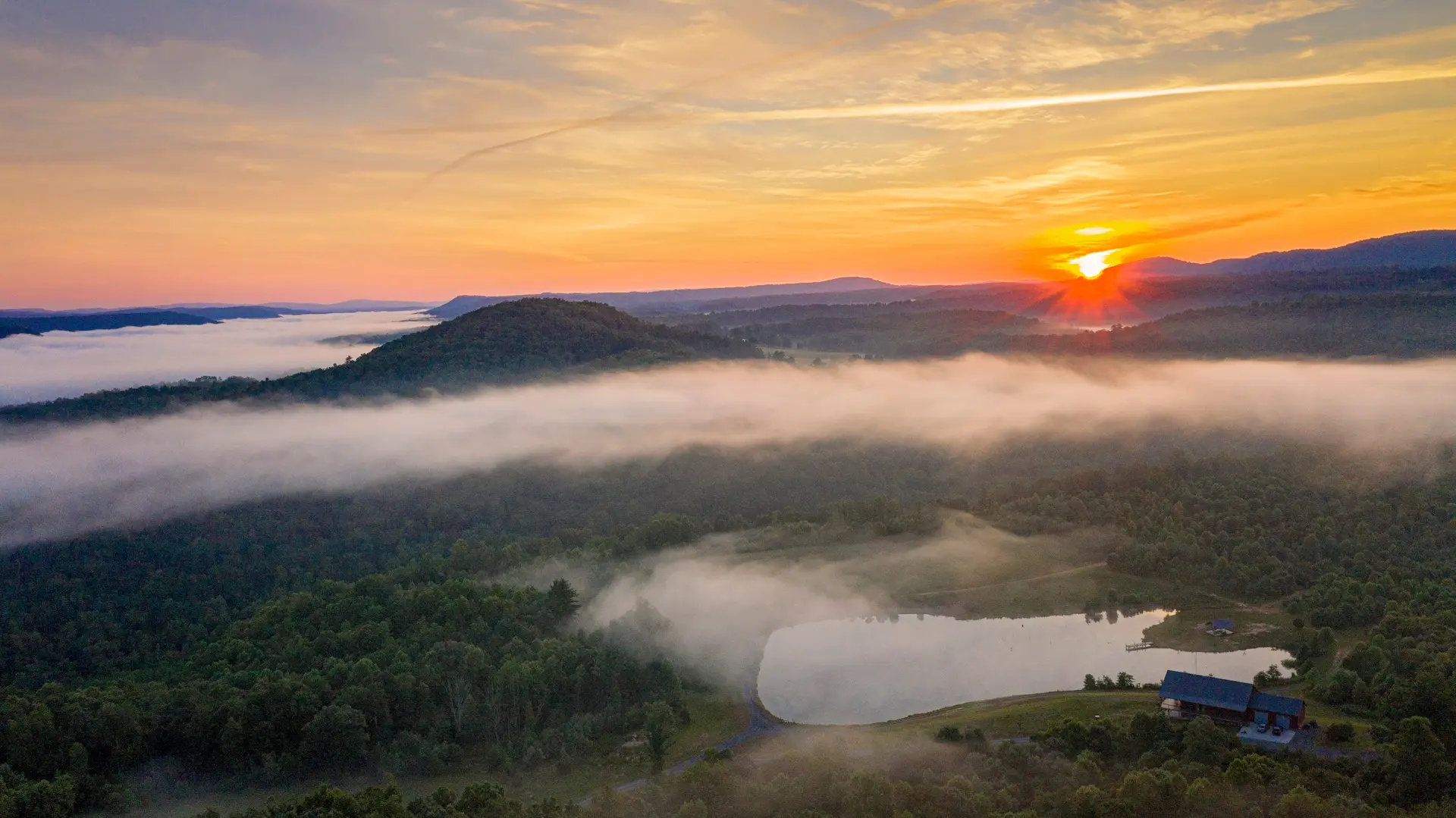 A breathtaking sunrise casts golden light over the misty mountains and rolling forested hills of Mercer County, WV, with fog drifting through the valleys and a serene pond reflecting the sky below.