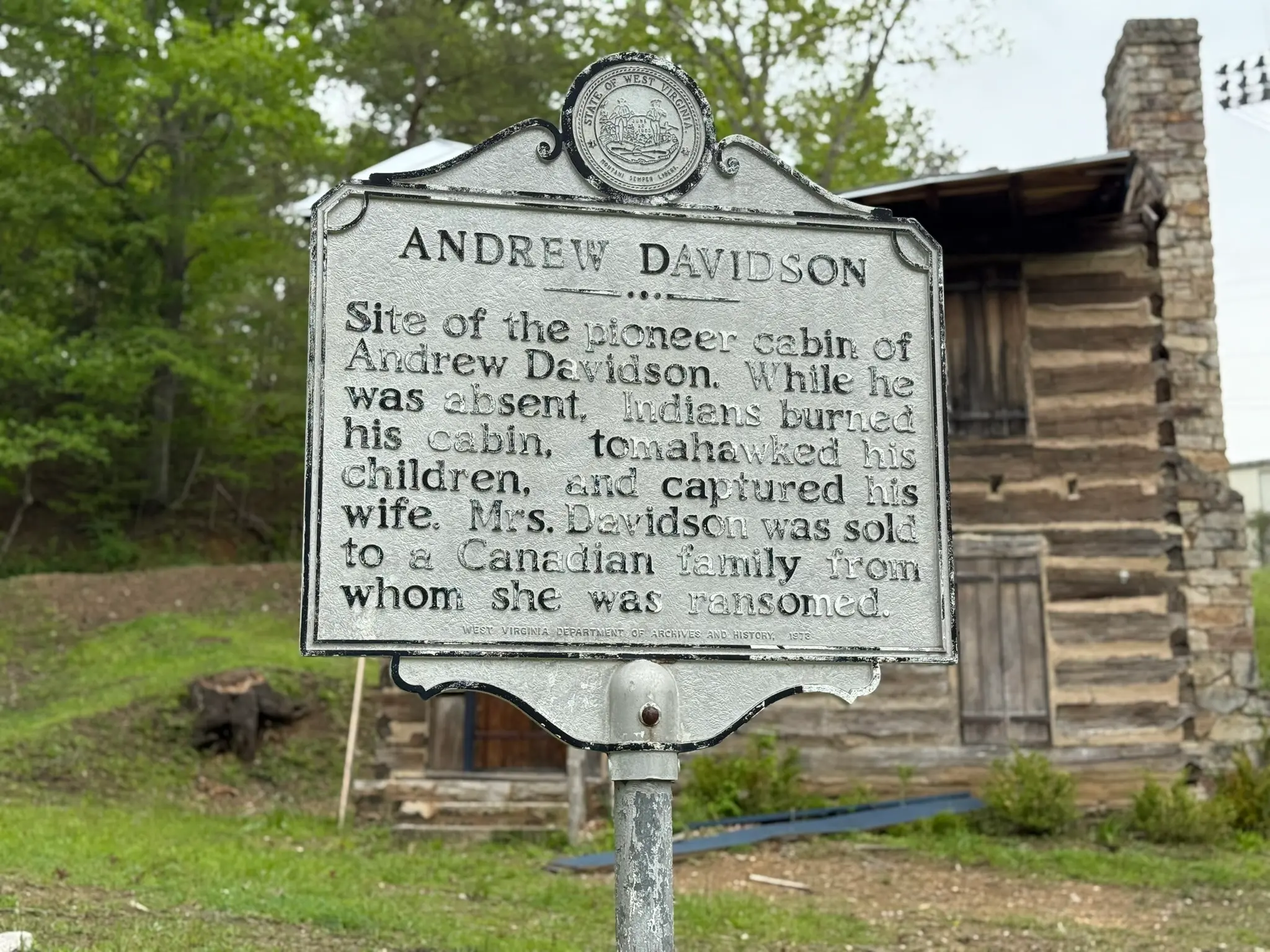 A historical marker in Mercer County, WV, stands in front of a preserved log cabin, detailing the site of Andrew Davidson’s pioneer home and its tragic past during early frontier conflicts.
