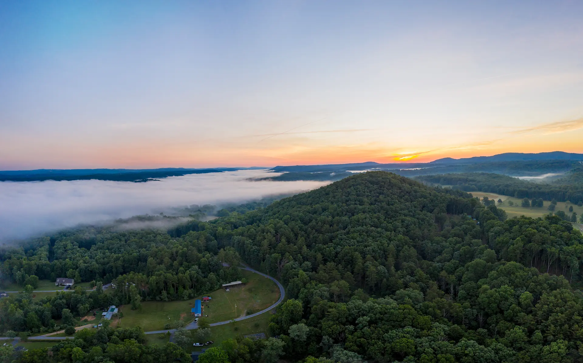 A wide aerial view of Mercer County, WV, at sunrise, showcasing tree-covered hills, rolling farmland, and low-lying morning fog stretching across the horizon under a glowing sky.