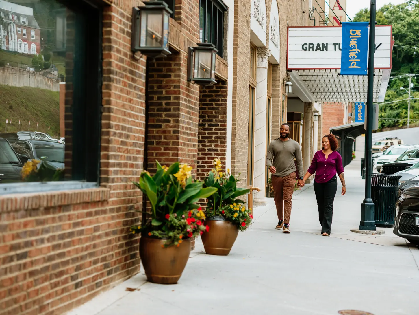 A couple strolls hand-in-hand down a charming sidewalk in downtown Bluefield, WV, passing colorful planters and the historic Granada Theater marquee.