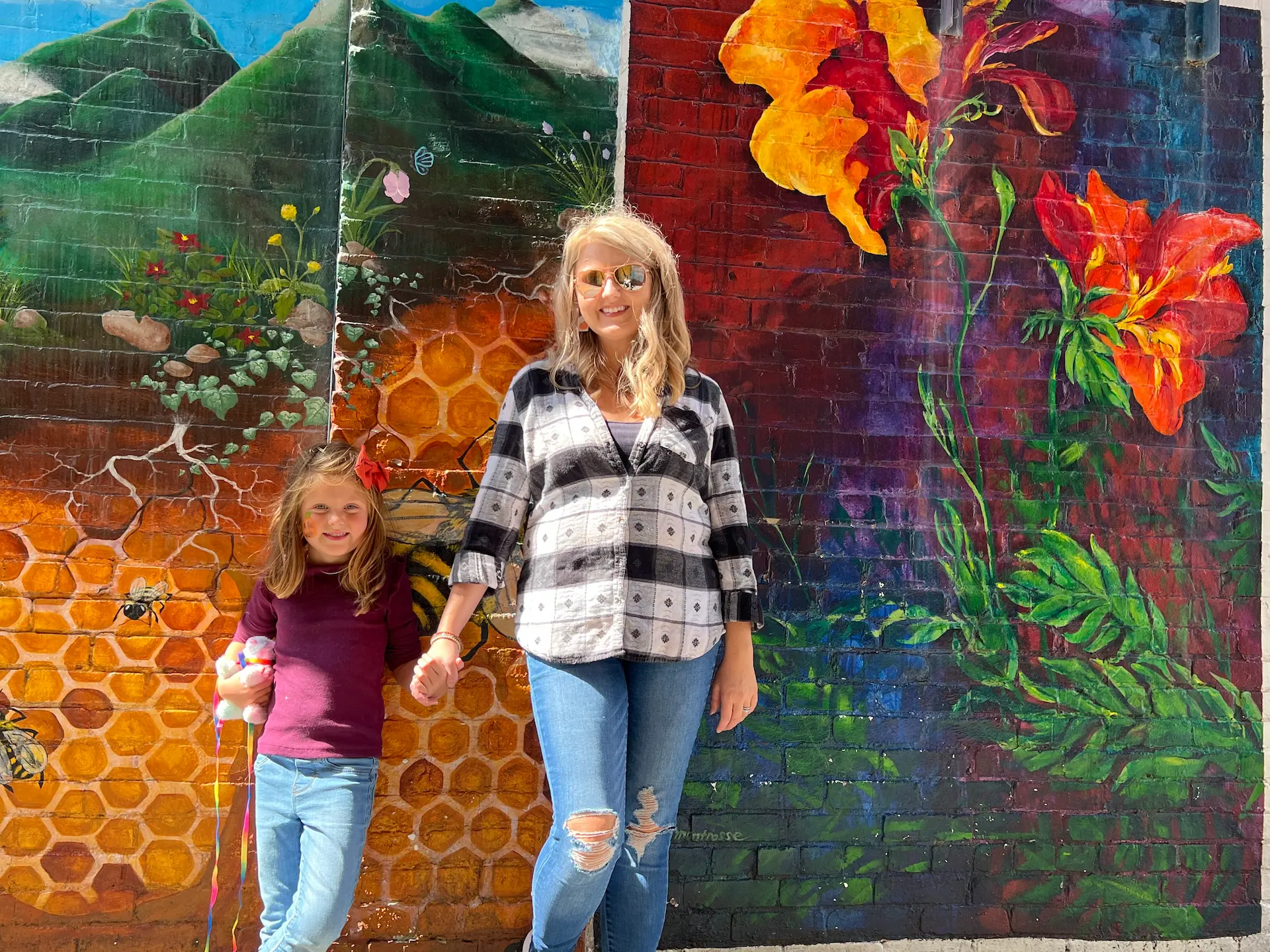 A woman and child smile in front of a colorful mural in Mercer County, WV, featuring vibrant flowers, honeycomb patterns, and scenic mountains.