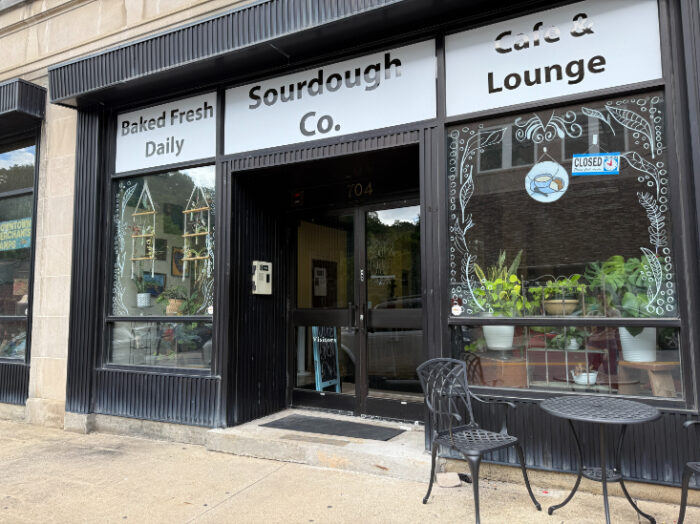 Entrance to Sourdough Co. Cafe, with large display windows trimmed in black, a bistro table, and plants in the windows.