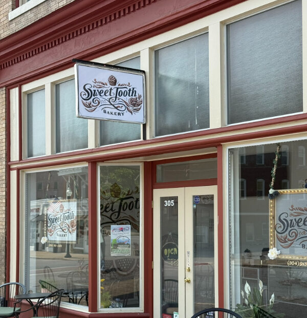 Storefront of Sweet Tooth Bakery, featuring large front windows, a glass door, and a hanging sign with the bakery's name and decorative design above the entrance.