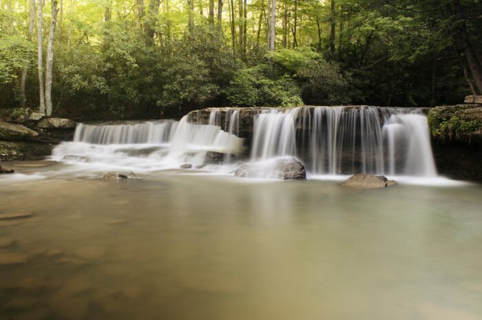A gentle waterfall at Mash Fork Falls in Mercer County, West Virginia, cascades over a wide rock ledge into a calm pool surrounded by lush green forest.