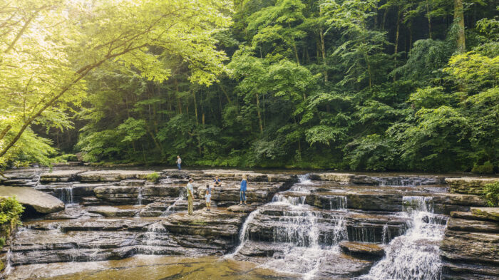 Visitors stand on the layered rock ledges of Campbell Falls at Camp Creek State Park in Mercer County, West Virginia. The 15-foot waterfall cascades in multiple streams over wide stone shelves, framed by lush green forest.