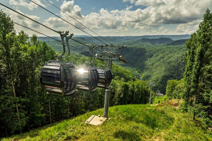 Glass-enclosed gondola cars riding above a lush, forested mountain landscape with rolling Appalachian hills stretching into the distance under a partly cloudy sky.