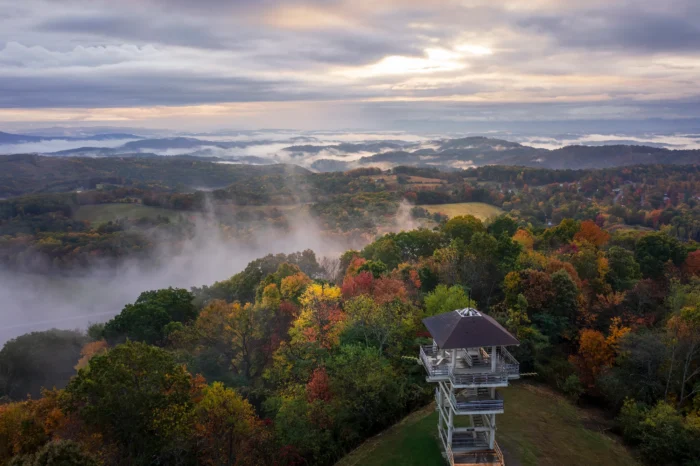 Aerial view of a wooden lookout tower surrounded by autumn foliage with misty mountains and dramatic clouds in the background.