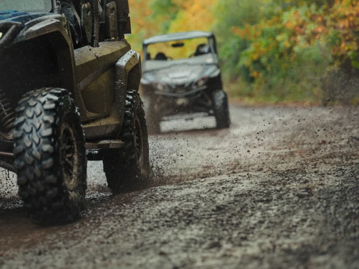 Muddy off-road vehicles driving on a dirt trail with autumn foliage in the background.