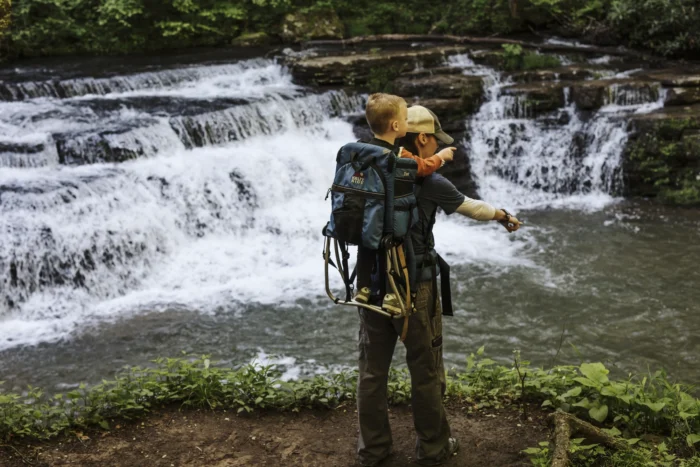 An adult carrying a child in a backpack carrier while standing near a cascading waterfall.